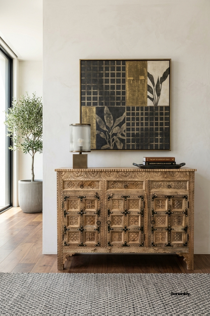 Hand-carved rustic 3-door credenza featuring paneled geometric relief carvings,
three upper utility drawers, and traditional dark iron strap and hinge accents.
Styled in an elegant foyer beneath modern mixed-media art with an olive tree
and fluted glass lamp, this artisan sideboard brings heritage warmth and statement storage.
