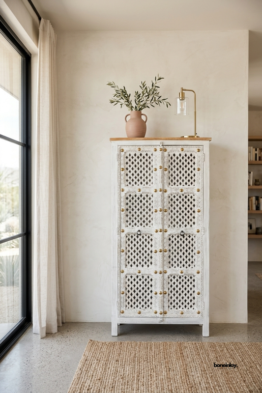 Hand-carved white distressed wood tall cabinet featuring lattice screen doors, decorative brass studs, and a natural wood top. Styled in a sunlit modern entryway next to floor-to-ceiling windows and a natural jute rug, this rustic armoire offers unique storage and artisan charm for luxury homes.