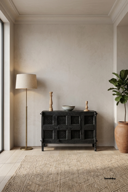 Artisan-crafted 2-door accent cabinet featuring traditional lattice fretwork door panels,
carved rosette border moldings, and antique metal latch hardware in a matte charcoal black finish.
Styled in a warm minimalist room beside a tall potted tree and brass floor lamp,
this compact credenza delivers dramatic rustic elegance and functional concealed storage.