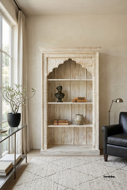 Handcrafted tall open bookshelf featuring a classic scalloped Jharokha arch crown,
fluted rope-carved pilasters, and four spacious display shelves in a distressed whitewash.
Styled in a cozy reading corner beside a black leather armchair and modern task lamp,
this artisan étagère adds regal character and practical storage to home libraries.