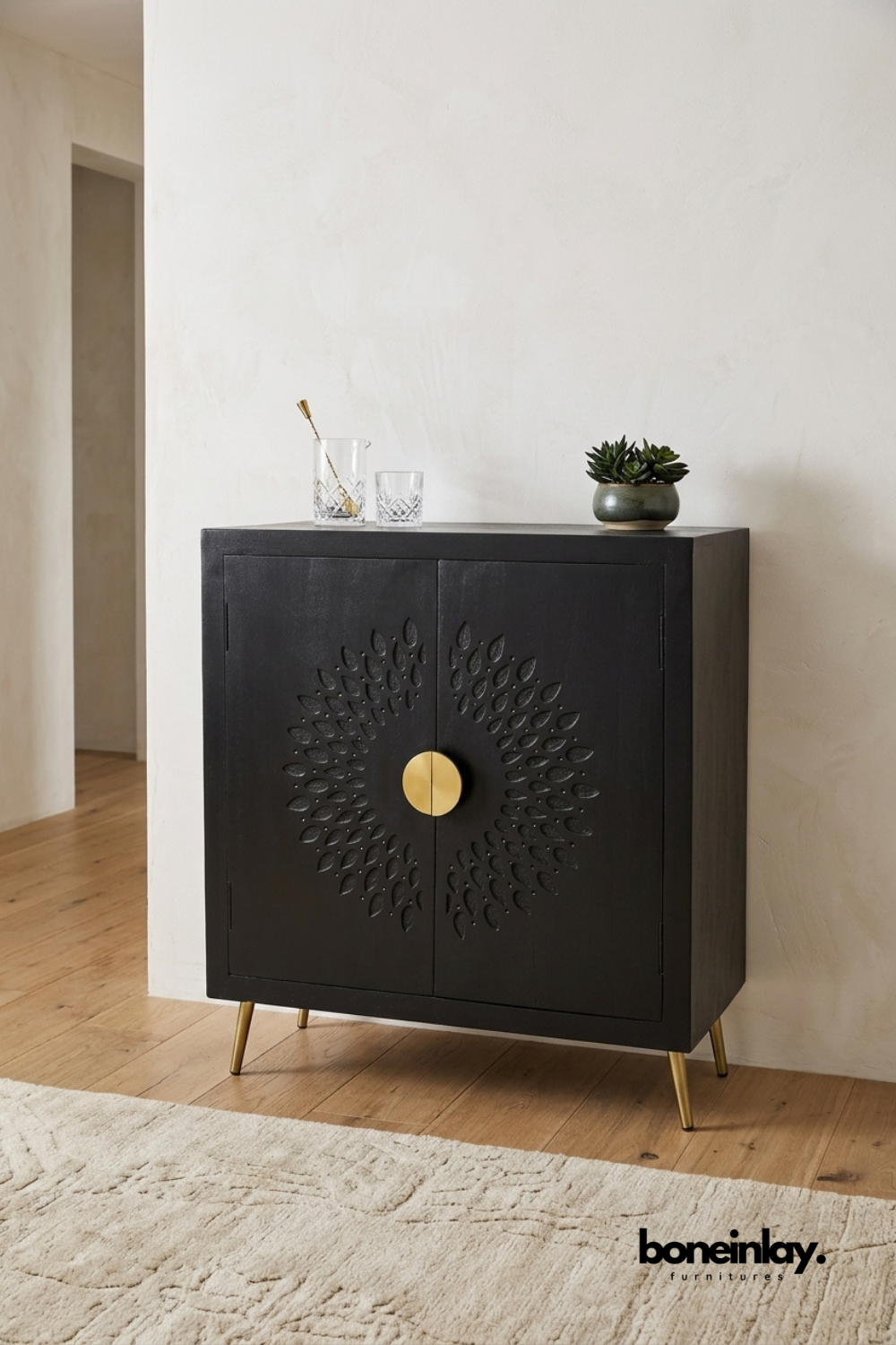 Matte black wood cabinet featuring a hand-carved sunburst leaf pattern radiating from a round brass knob. Slim tapered brass legs add a refined mid-century modern silhouette to the design. Styled with crystal glassware, a brass bar spoon, and a small potted succulent. A sophisticated bar cabinet or accent piece for modern living and entertaining spaces.