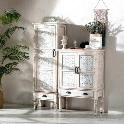 Hand-carved wood cabinet with a distressed white door and lower drawer, styled in a bright room with plants.