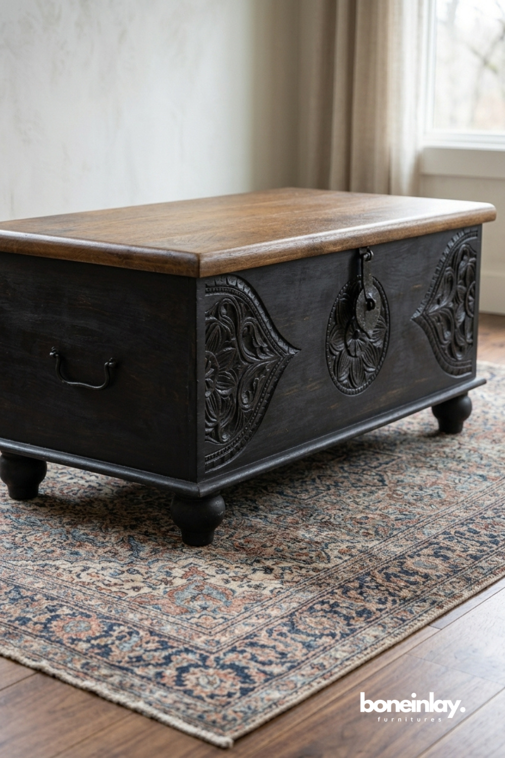 Black carved wood trunk with a natural wood tabletop.
Front panel features hand-carved medallion and floral corner details.
Round bun feet and a wrought iron latch add rustic character.
Styled on a patterned rug as a coffee table or storage bench.
