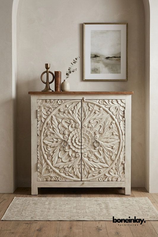 Whitewashed hand-carved wood accent cabinet featuring a detailed floral medallion pattern on double doors. Topped with a natural wood surface and finished with round bun feet for rustic charm. Styled with a brass candlestick, vintage book, and eucalyptus sprig beneath framed art. A charming accent cabinet for boho and farmhouse-inspired interiors.