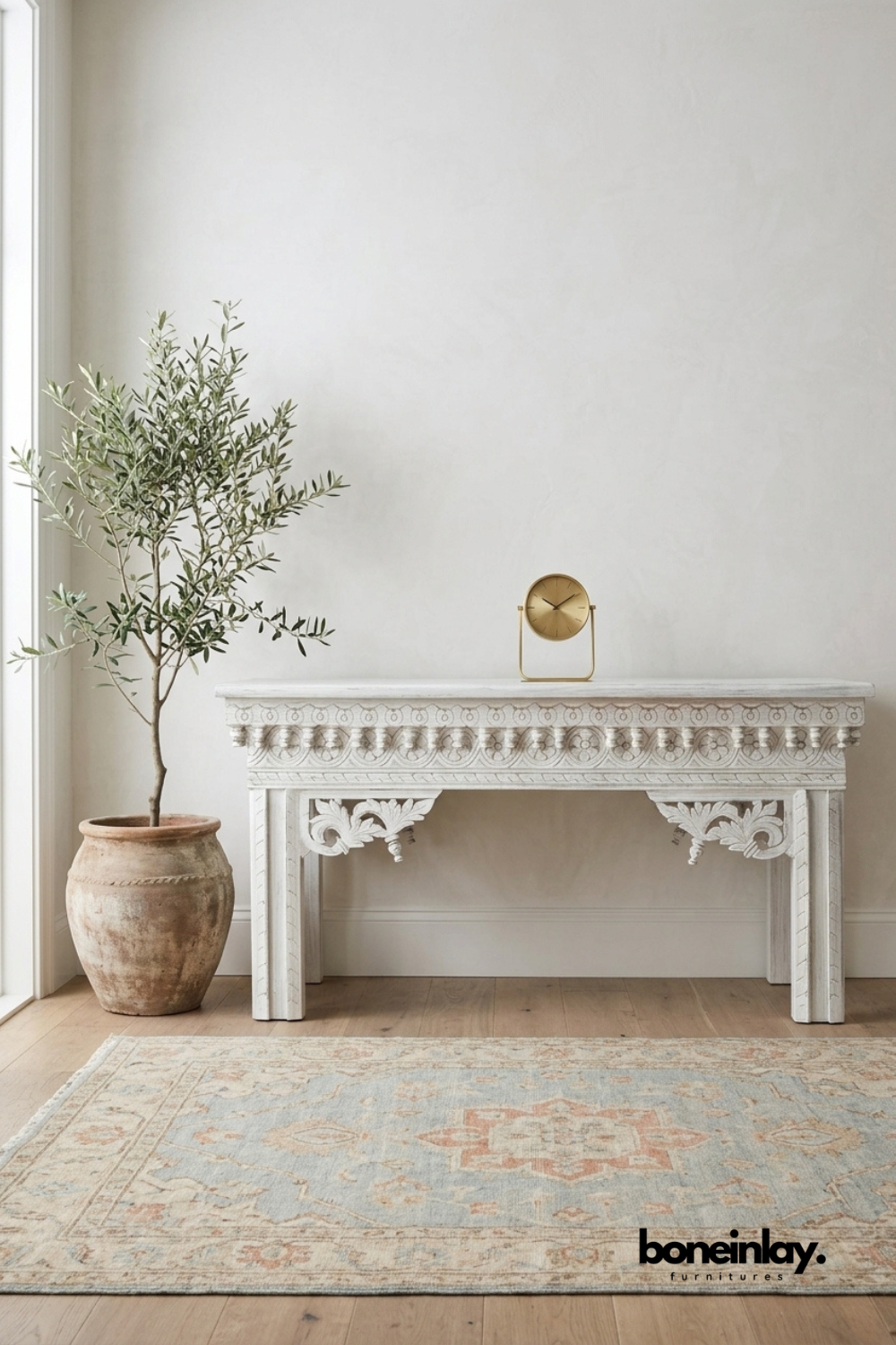 Whitewashed carved wood console table featuring ornate scalloped trim and floral scroll leg brackets. Traditional beaded and rosette detailing runs along the apron for a vintage European-inspired look. Styled with a terracotta planter, olive tree, and brass table clock on a Persian-style rug. An elegant statement console for hallways, entryways, and living room walls.