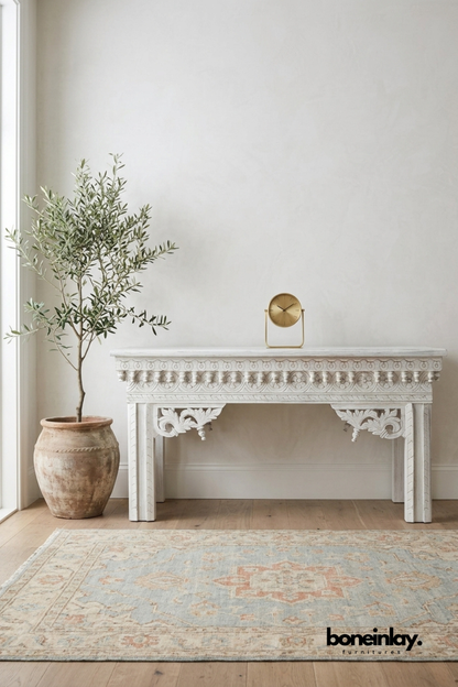 Whitewashed carved wood console table featuring ornate scalloped trim and floral scroll leg brackets. Traditional beaded and rosette detailing runs along the apron for a vintage European-inspired look. Styled with a terracotta planter, olive tree, and brass table clock on a Persian-style rug. An elegant statement console for hallways, entryways, and living room walls.