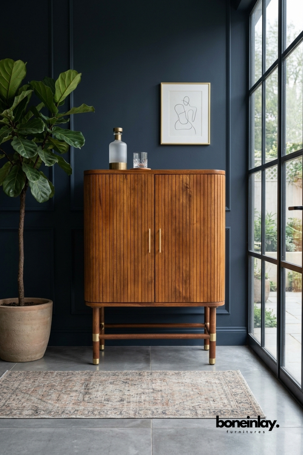Warm walnut wood bar cabinet featuring a rounded fluted silhouette with vertical ribbed detailing on double doors. Brass pull handles and brass-capped legs add refined metallic contrast to the rich wood tone. Styled with a glass decanter and crystal tumbler against a deep navy paneled wall. A sophisticated bar cabinet for dining rooms and entertaining spaces.