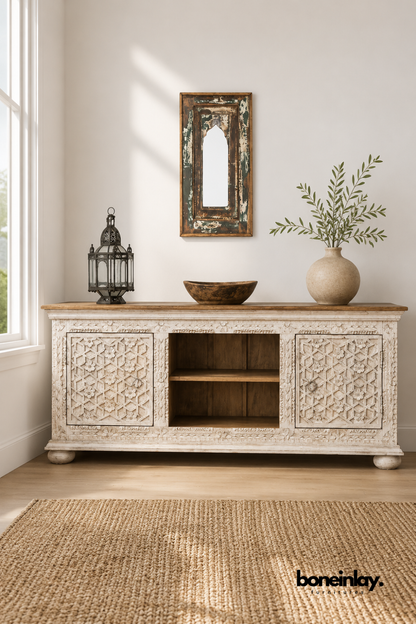 Whitewashed carved wood TV console with a star lattice door pattern.
Open center shelving sits between two intricately carved cabinet doors.
Natural wood tabletop contrasts with the whitewashed carved base.
Styled beneath a rustic arched mirror and wooden bowl.