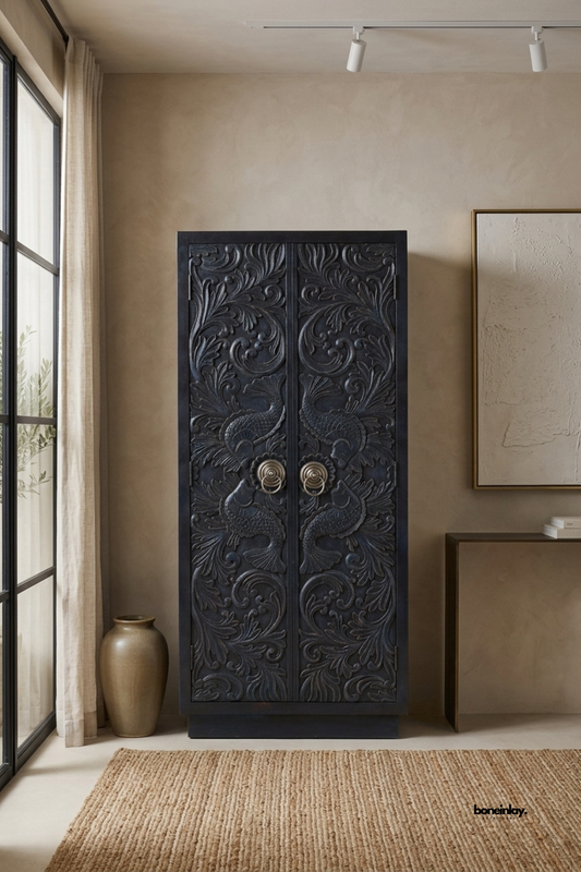 Artisan hand-carved solid wood tall armoire cabinet featuring deep relief botanical scrollwork, stylized fish motifs, and vintage brass ring pulls in an antique charcoal black finish. Styled on a woven jute rug beside a tall black window frame, this statement linen storage cabinet delivers heirloom artistry and dramatic grandeur to luxury spaces.