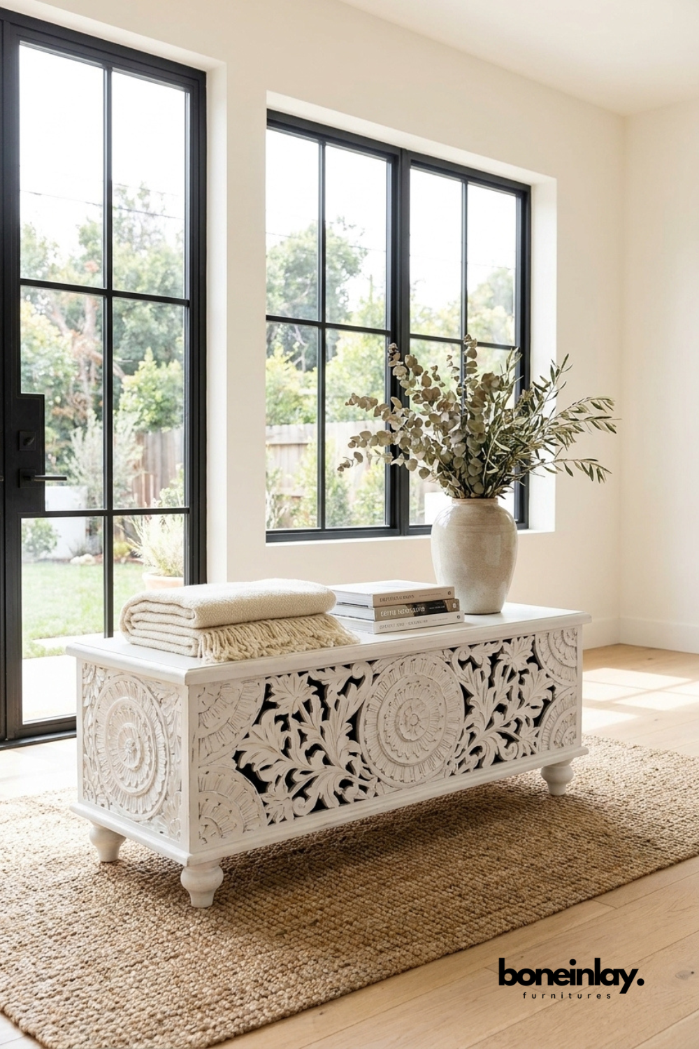 Hand-carved white storage chest styled with decorative books and a ceramic vase, placed in a bright modern room with large black-framed windows and natural light.