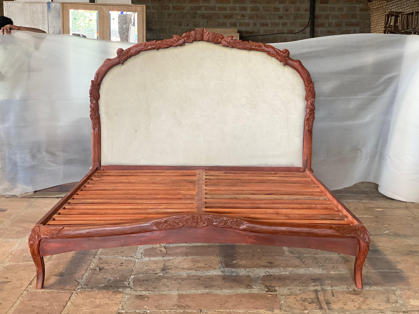 Unfinished walnut-stained Menagerie bed frame in solid mango wood,
arched upholstered headboard panel with hand-carved floral border,
slatted base and cabriole legs shown in artisan workshop setting.