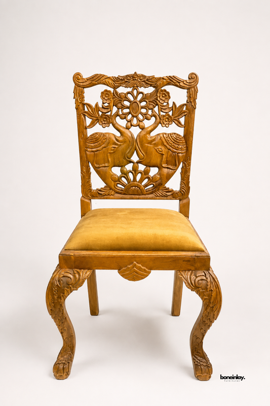 Hand-carved solid teak wood accent chair showcasing an ornate backrest with facing royal elephant motifs, floral scroll relief work, cabriole legs, and a rich ochre gold velvet upholstered seat cushion. Displayed against a clean studio backdrop, this artisan dining chair brings regal Indian heritage craftsmanship and classic luxury to dining rooms.