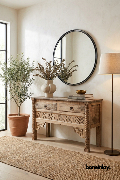 Hand-carved wood console table with a distressed natural finish.
Two front drawers sit above a carved jaali lattice panel.
Spiral-turned front legs add traditional artisanal detailing.
Styled beneath a round black-framed mirror in an entryway.