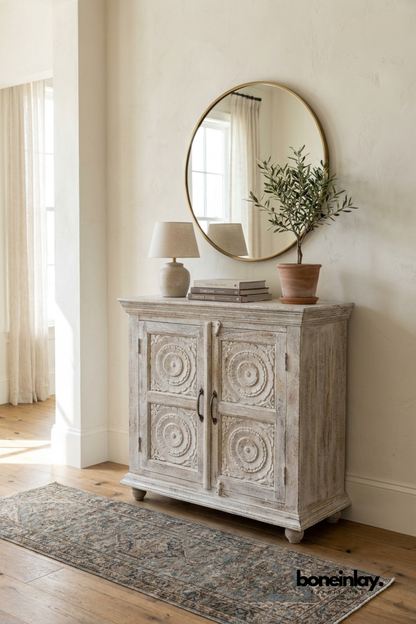 Whitewashed carved wood cabinet with round medallion door panels.
Detailed circular rosette carving repeats across all four doors.
Turned bun feet and a molded top edge add classic character.
Styled beneath a round brass mirror on a vintage-style runner rug.
