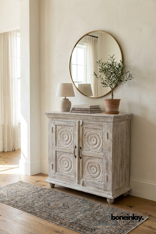 Whitewashed carved wood cabinet with round medallion door panels.
Detailed circular rosette carving repeats across all four doors.
Turned bun feet and a molded top edge add classic character.
Styled beneath a round brass mirror on a vintage-style runner rug.