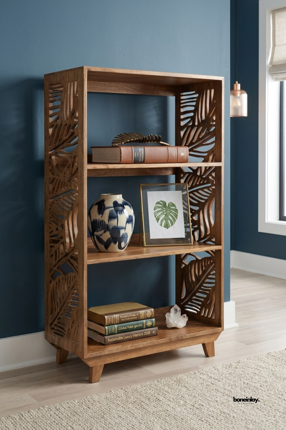 Walnut wood bookshelf featuring intricate palm leaf cutout side panels and three open display shelves. Slim tapered legs create a light, sculptural silhouette against a deep blue accent wall. Styled with a hand-painted vase, framed botanical print, stacked books, and a crystal cluster. A striking open bookshelf for living rooms and reading nook styling.