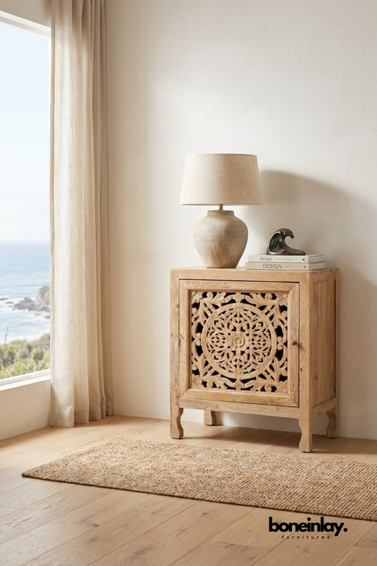 Hand-carved wooden cabinet with intricate floral detailing styled beside a large window, featuring a ceramic table lamp, decorative books, and coastal-inspired neutral decor.