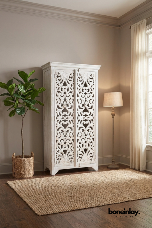 Whitewashed carved wood armoire with an intricate open scroll lattice front.
Flowing vine and heart-shaped carving spans both door panels.
Crown molding top and angled feet complete the elegant silhouette.
Styled beside a woven basket planter and antique brass floor lamp.