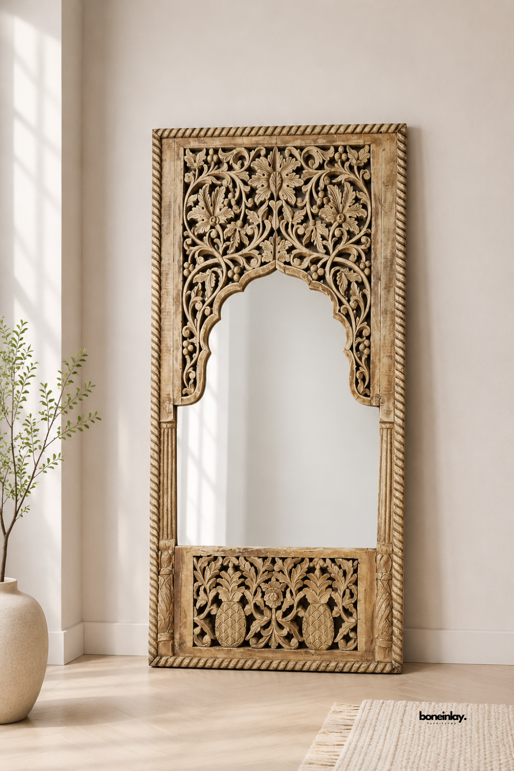 Hand-carved wooden full-length floor mirror featuring ornate botanical relief work, pineapple motifs, and an elegant scalloped arch mirror cutout. Leaning in a sunlit neutral hallway next to a ceramic vase and woven rug, this rustic artisan accent mirror brings timeless bohemian elegance and warmth to luxury home decor.