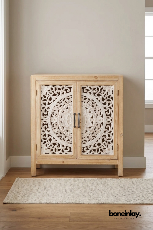 Whitewashed wood accent cabinet with a hand-carved medallion pattern.
Two doors feature intricate scroll and flower carving in a circular motif.
Simple block legs give it a light, understated base.
Styled with a stack of books in front of an interior doorway.