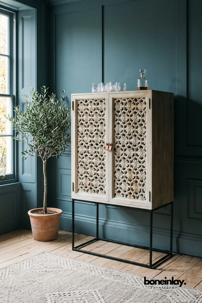 Hand-carved wood bar cabinet with an intricate floral lattice design.
Two carved doors open to reveal storage for glassware and bottles.
Raised on a slim black iron stand for a modern-meets-vintage look.
Styled against a deep teal wall with an olive tree beside it.