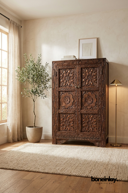 Hand-carved brown wood armoire cabinet with a rich walnut tone.
Floral medallion carvings cover both cabinet door fronts.
Ornate vine and border detailing frames the entire piece.
Styled beside an olive tree and brass floor lamp.