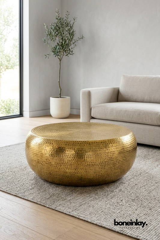 Round hammered brass coffee table with a rich gold finish and rounded drum silhouette. Placed on a woven jute rug beside a linen sofa and potted olive tree. Artisanal hammered texture adds handcrafted warmth and sculptural presence to the space. A statement coffee table combining Old World craftsmanship with modern living room styling.