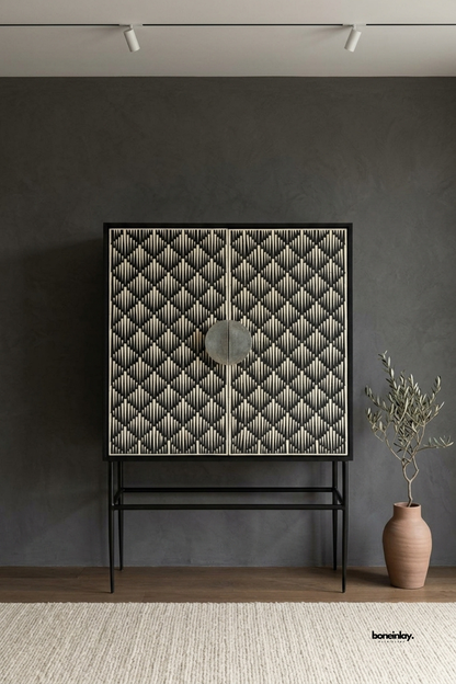 Handcrafted bone inlay tall bar cabinet featuring a black and white geometric optical pattern, two tall doors, circular metal handle pulls, and a slender black iron base. Styled against a dark charcoal accent wall with a potted olive tree and woven rug, this artisan statement armoire delivers modern luxury and sophisticated concealed storage.