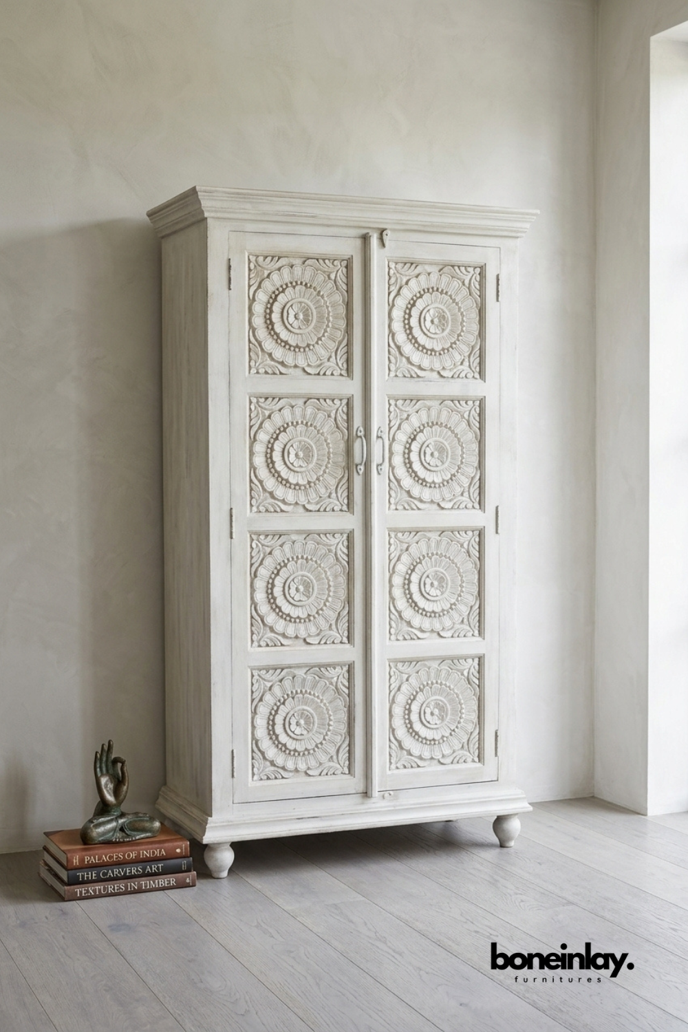 White carved wood wardrobe with a repeating floral medallion pattern.
Four carved panels run the full height of both cabinet doors.
Crown top molding and round wood feet complete the traditional design.
Styled beside a stack of books and a small bronze figurine.