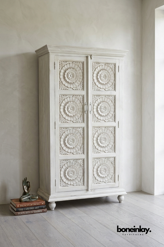 White carved wood wardrobe with a repeating floral medallion pattern.
Four carved panels run the full height of both cabinet doors.
Crown top molding and round wood feet complete the traditional design.
Styled beside a stack of books and a small bronze figurine.