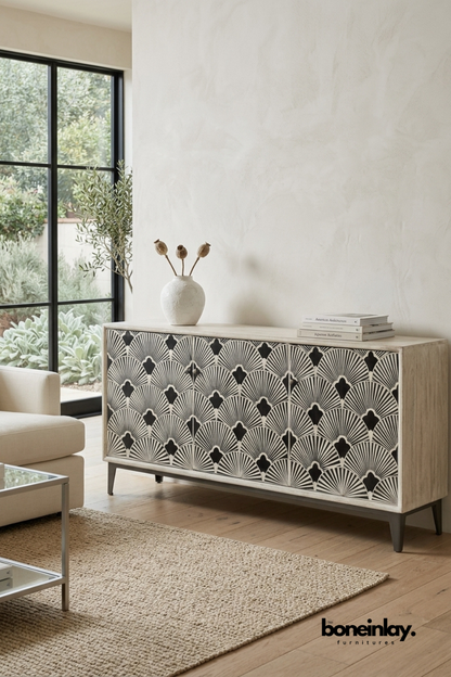 Natural wood media unit featuring a black and ivory scalloped fan bone inlay pattern across three doors. Finished with tapered dark wood legs for a warm mid-century inspired look. Styled with a ceramic vase, dried botanicals, and stacked books near a glass coffee table. An eye-catching storage piece for living and dining room settings.