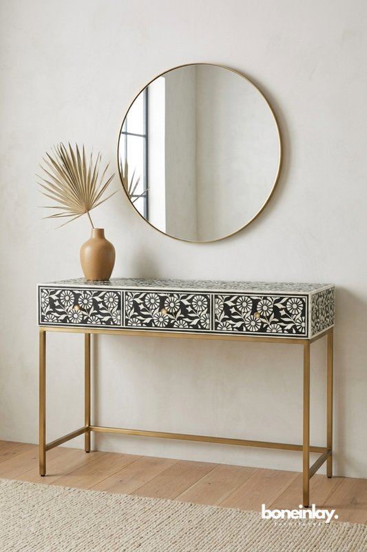 Black and white bone inlay console table featuring a bold floral daisy pattern across the drawer front. Finished with brass drawer knobs and a sleek gold metal frame base. Styled with a round brass-framed mirror and a mustard ceramic vase with a dried palm frond. A striking entryway console blending graphic pattern with modern minimalist lines.