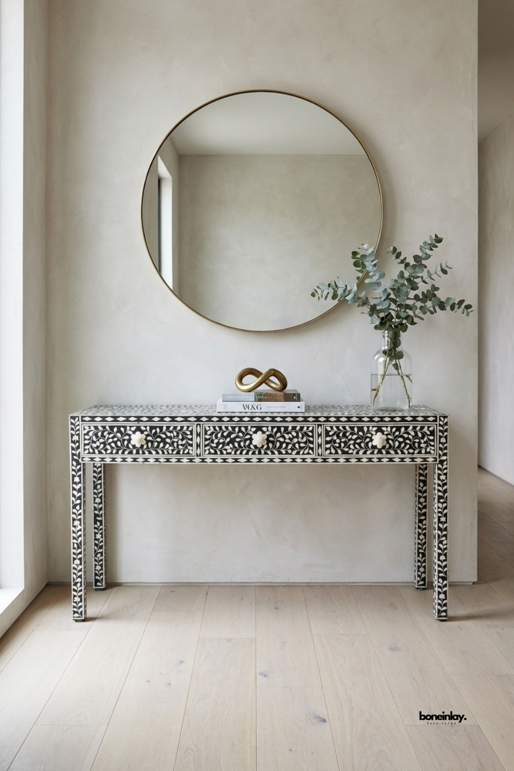 Black and white bone inlay console table featuring a delicate floral vine pattern across three drawers and tapered legs. Bone flower-shaped knobs add a refined handcrafted detail to the graphic design. Styled beneath a round brass-framed mirror with a glass vase of eucalyptus and a brass infinity sculpture. An elegant statement console for entryways and hallway styling.
