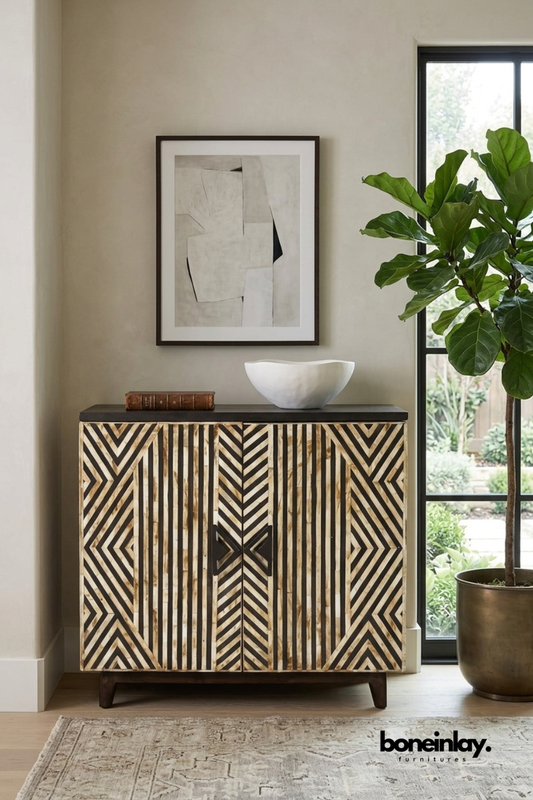 Black and ivory bone inlay accent cabinet featuring a bold chevron stripe pattern on double doors. Finished with a dark wood top, angled legs, and a sculptural X-shaped door pull. Styled with a leather-bound book and organic white bowl beneath framed abstract art. A graphic statement cabinet for modern eclectic interiors.