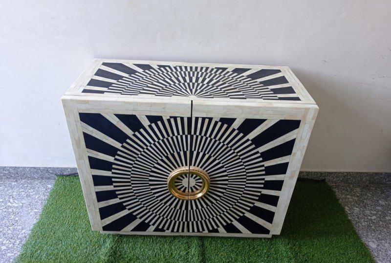Decorative black and white bone inlay bar cabinet featuring a geometric sunburst pattern across the two-door front. Its circular gold-finished handles provide a bold metallic accent against the high-contrast black resin and ivory bone design. The handcrafted cabinet adds statement storage to a home bar, living room, or maximalist interior and is intended for indoor use.