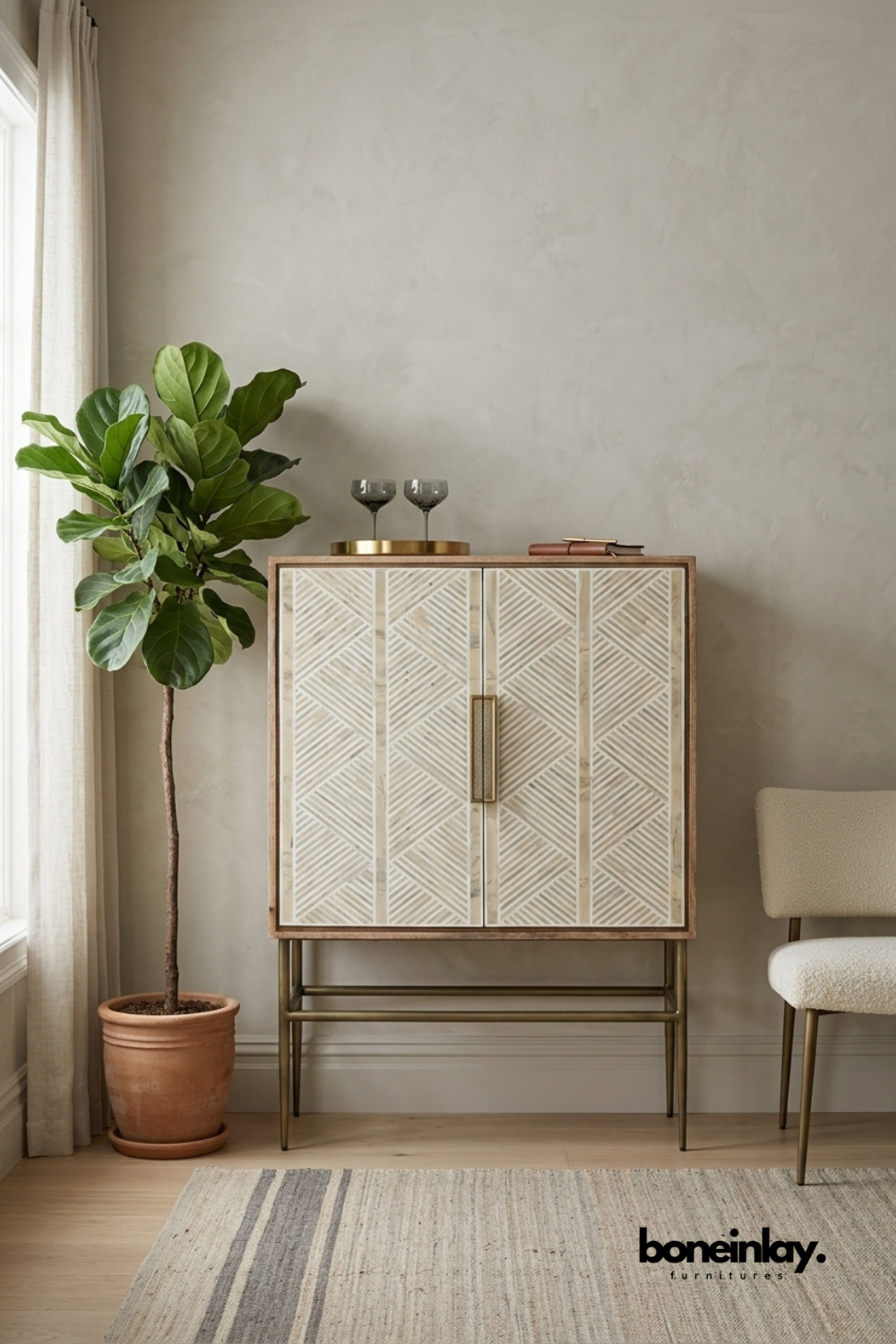Natural wood bar cabinet featuring an ivory bone inlay chevron pattern across double doors with a brass pull handle. Slim brass legs and stretcher base add a refined mid-century modern silhouette. Styled with etched glassware, a brass tray, and a leather journal beside a fiddle leaf fig. A sophisticated bar cabinet for entertaining spaces and living rooms.