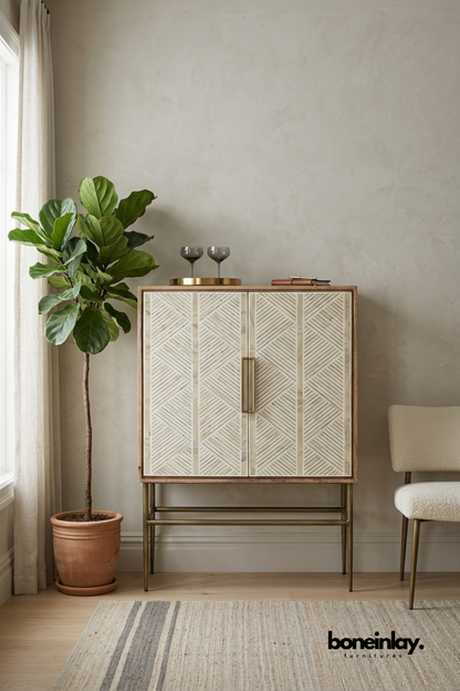 Natural wood bar cabinet featuring an ivory bone inlay chevron pattern across double doors with a brass pull handle. Slim brass legs and stretcher base add a refined mid-century modern silhouette. Styled with etched glassware, a brass tray, and a leather journal beside a fiddle leaf fig. A sophisticated bar cabinet for entertaining spaces and living rooms.