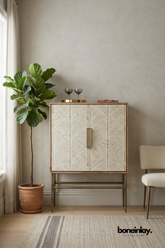 Natural wood bar cabinet featuring an ivory bone inlay chevron pattern across double doors with a brass pull handle. Slim brass legs and stretcher base add a refined mid-century modern silhouette. Styled with etched glassware, a brass tray, and a leather journal beside a fiddle leaf fig. A sophisticated bar cabinet for entertaining spaces and living rooms.