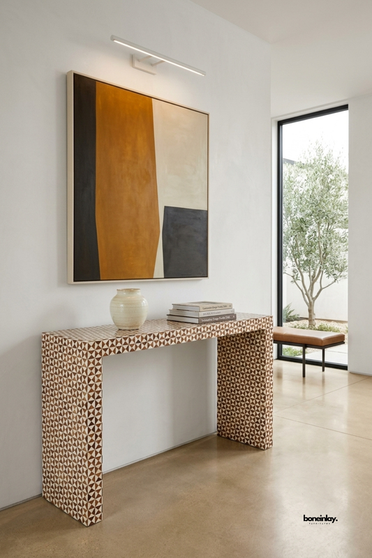 Brown and ivory bone inlay console table featuring a dense geometric triangle mosaic pattern across the entire surface. Clean waterfall-style legs create a sleek architectural silhouette in warm earthy tones. Styled with a ceramic vase and stacked books beneath a bold abstract painting near a garden view. A striking statement console for entryways and hallway styling.