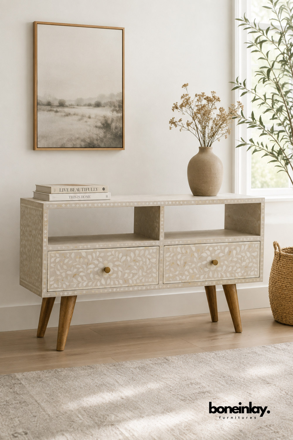 Cream bone inlay TV console with a delicate leaf vine pattern.
Two open display shelves sit above a pair of drawers with brass knobs.
Angled wood legs give the piece a light mid-century stance.
Styled beneath a framed landscape print and a stoneware vase.