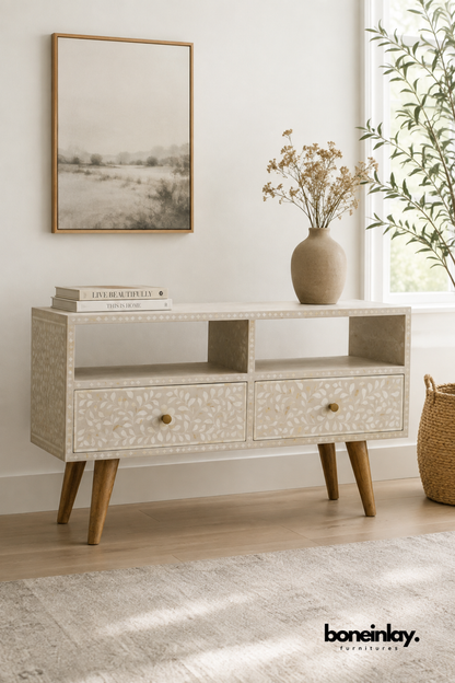 Cream bone inlay TV console with a delicate leaf vine pattern.
Two open display shelves sit above a pair of drawers with brass knobs.
Angled wood legs give the piece a light mid-century stance.
Styled beneath a framed landscape print and a stoneware vase.