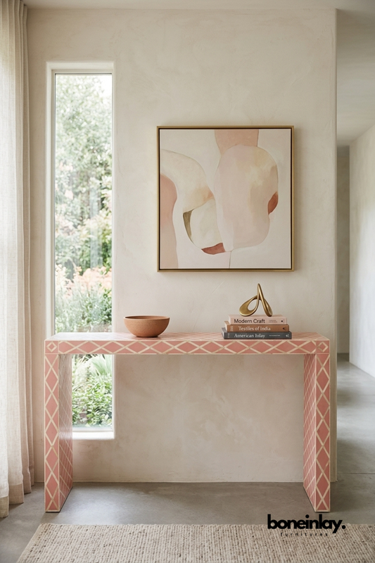 Blush pink bone inlay console table featuring a diamond trellis pattern with contrasting ivory detailing. Sleek waterfall-style legs create a clean architectural silhouette in soft pastel tones. Styled with a terracotta bowl, stacked books, and a brass sculpture beneath framed abstract art. A romantic statement console for entryways and feminine-inspired interiors.