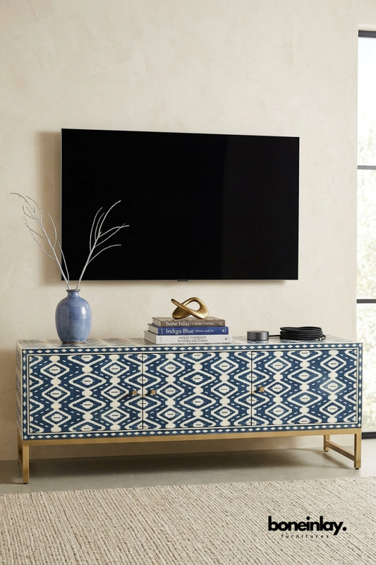 Navy blue and ivory bone inlay TV console featuring a bold ikat diamond pattern across three cabinet doors. Brass hardware and a slim gold metal base add refined contrast to the rich pattern. Styled beneath a wall-mounted television with a ceramic vase, books, and a smart speaker. A striking media console for eclectic and globally-inspired living rooms.