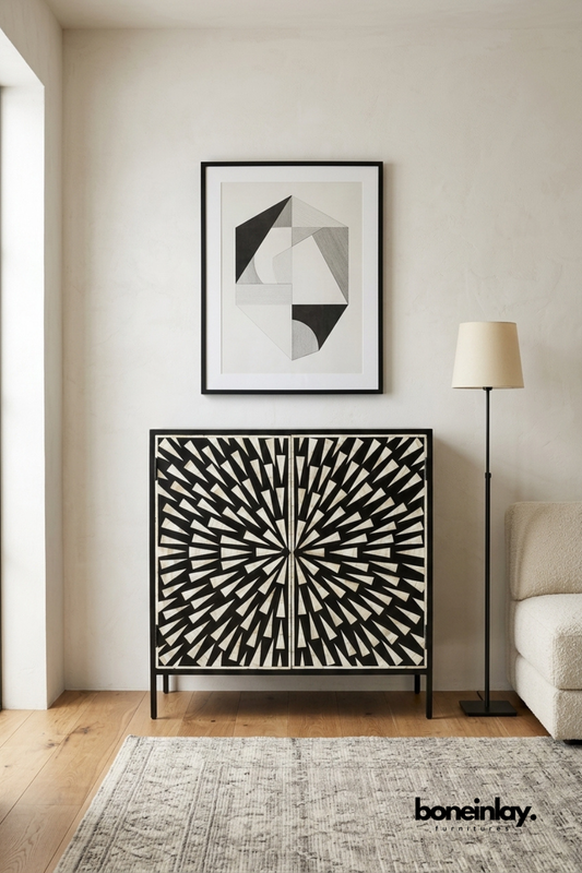 Black and white bone inlay accent cabinet featuring a radiating sunburst geometric pattern on double doors. Set on a slim black metal frame base for a modern architectural silhouette. Styled beneath framed abstract art beside a boucle sofa and floor lamp. A bold graphic statement cabinet for contemporary living room decor.