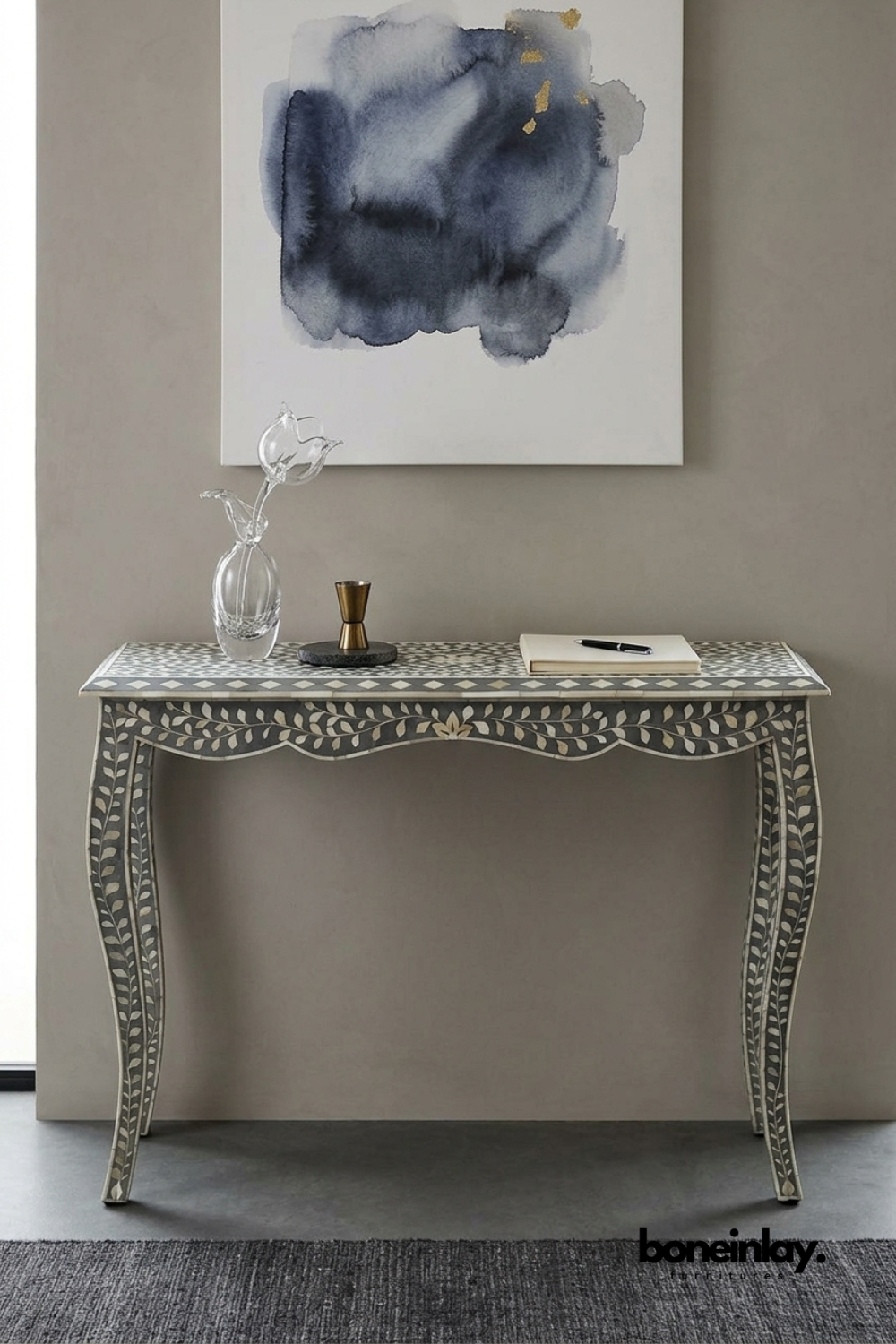 Grey and ivory bone inlay console table featuring a delicate floral vine pattern and scalloped skirt detailing. Elegant curved cabriole legs with matching leaf inlay complete the sophisticated silhouette. Styled with a glass tulip sculpture, brass jigger, and notebook beneath a watercolor painting. A refined statement console for entryways and hallway styling.
