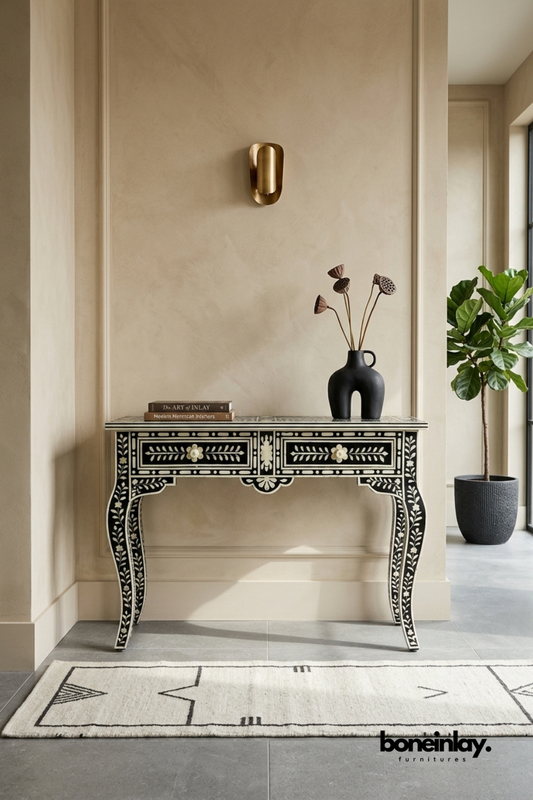 Black and white bone inlay console table featuring a delicate floral vine pattern and scalloped apron detailing. Elegant cabriole legs add a traditional, sculptural silhouette to the design. Styled with a black ceramic vase, dried lotus pods, and stacked books beneath a brass wall sconce. A sophisticated statement console for entryways and formal living spaces.