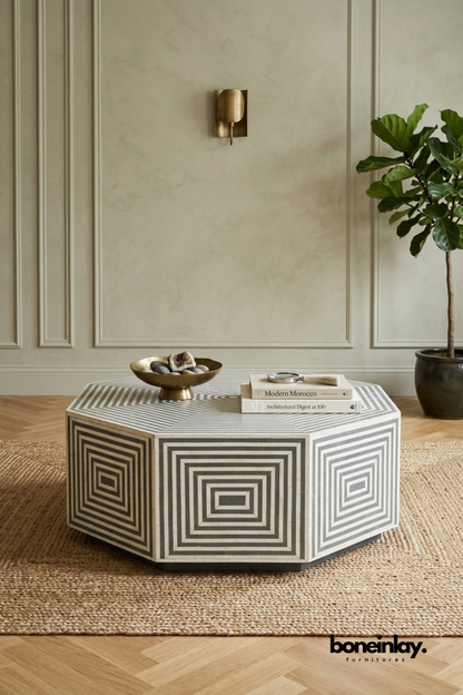 Black and white bone inlay coffee table featuring a bold octagonal silhouette with concentric geometric patterning. Striking symmetrical design wraps every angled panel for a dramatic visual centerpiece. Styled with a brass bowl of stones and stacked design books on a jute rug. An eye-catching statement coffee table for modern and eclectic living rooms.