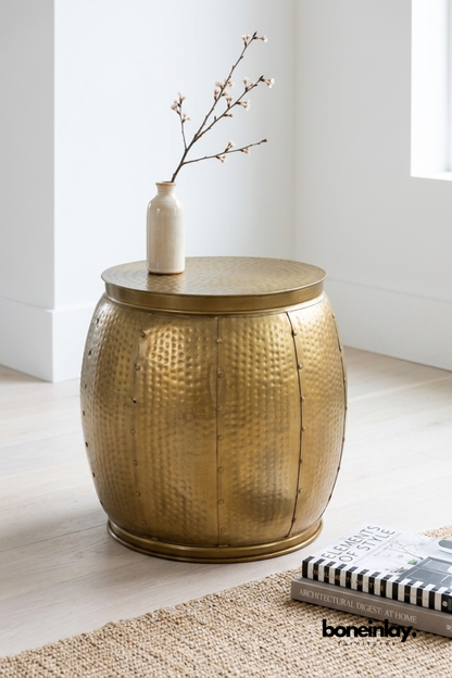 Hammered brass drum side table featuring a riveted barrel silhouette with a warm antique gold finish. Textured hammered detailing and visible seam rivets add artisanal, industrial character. Styled with a ceramic bud vase and flowering branch on a jute rug beside stacked books. A versatile accent table for living rooms, bedrooms, and reading nooks.