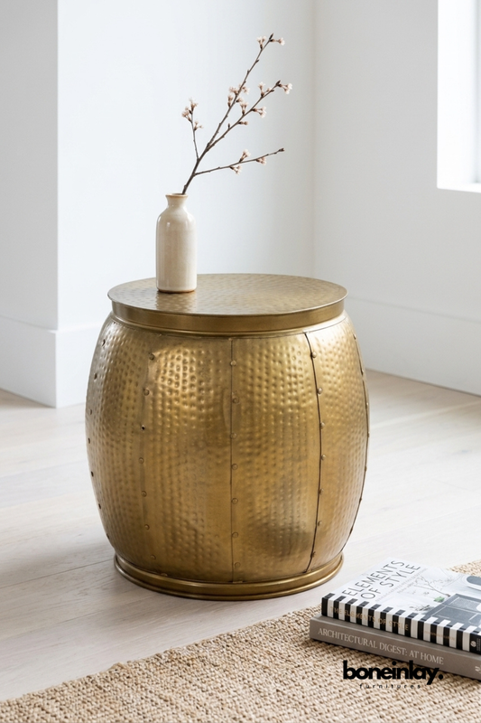 Hammered brass drum side table featuring a riveted barrel silhouette with a warm antique gold finish. Textured hammered detailing and visible seam rivets add artisanal, industrial character. Styled with a ceramic bud vase and flowering branch on a jute rug beside stacked books. A versatile accent table for living rooms, bedrooms, and reading nooks.
