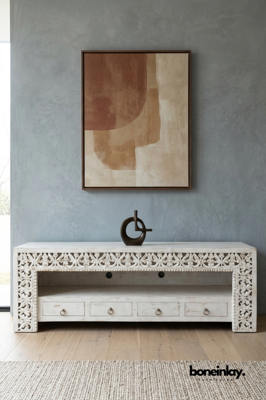White carved wood TV console with an open center display shelf.
Scroll and leaf carving frames the top edge and side panels.
Four lower drawers with ring pulls provide additional storage.
Styled beneath a warm-toned abstract art print on a grey wall.